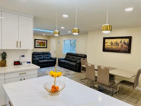 Open-plan interior with a white kitchen island in the foreground, dining table and chairs to the right, and two dark sofas in the background.