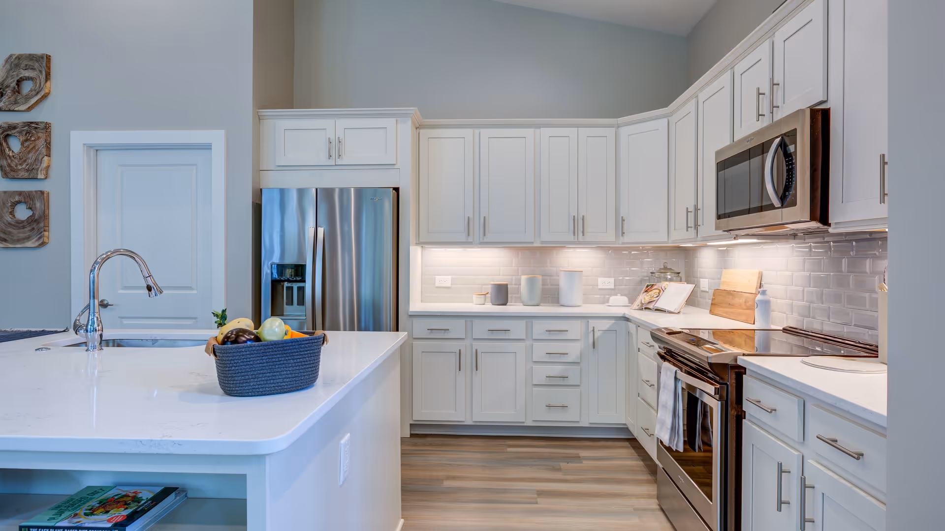 Modern kitchen with white cabinets, stainless steel refrigerator, oven, and microwave. A white island countertop with a sink and a basket of fruit is in the foreground. The kitchen has a light gray subway tile backsplash and wood flooring.