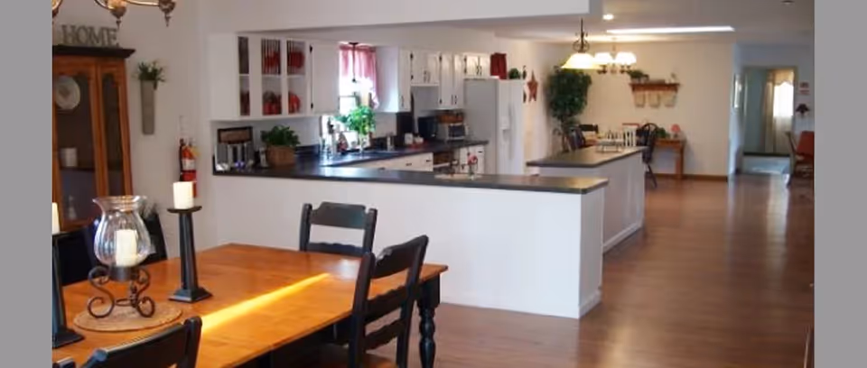 Interior view of a senior living facility dining and kitchen area with wooden floors, a wooden dining table with black chairs, candle holders on the table, white kitchen cabinets, a refrigerator, and a countertop separating the kitchen from the dining space. There are plants and decorative items on the counters and shelves.