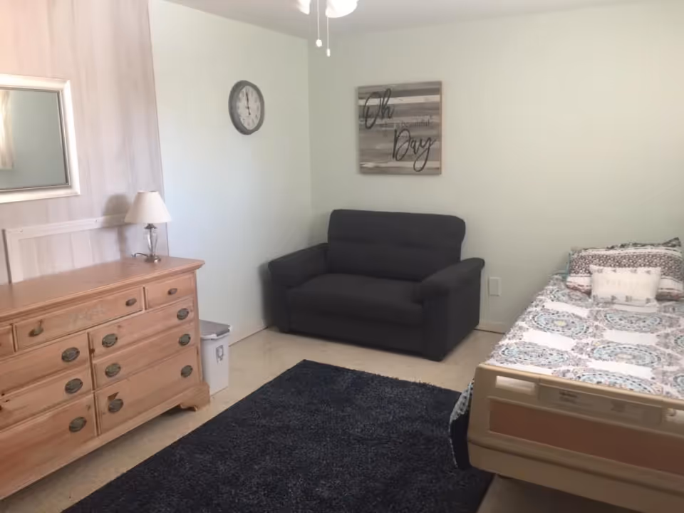 A small bedroom with a wooden dresser and lamp, a dark loveseat, and a single bed with patterned bedding.