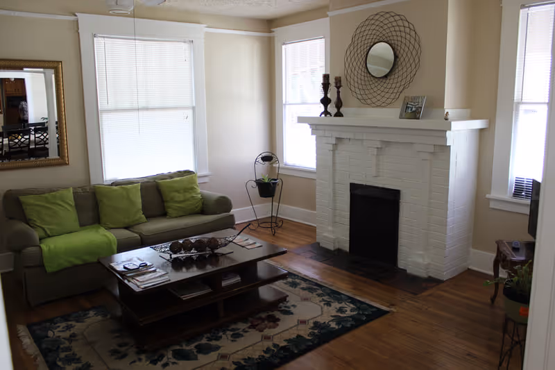 A cozy living room with a green sofa adorned with green cushions and a green throw blanket. In front of the sofa is a wooden coffee table with decorative items and magazines. The room features a white brick fireplace with a round decorative mirror and candle holders on the mantel. There are two windows with blinds, a patterned area rug on the wooden floor, and a few small plants placed around the room.