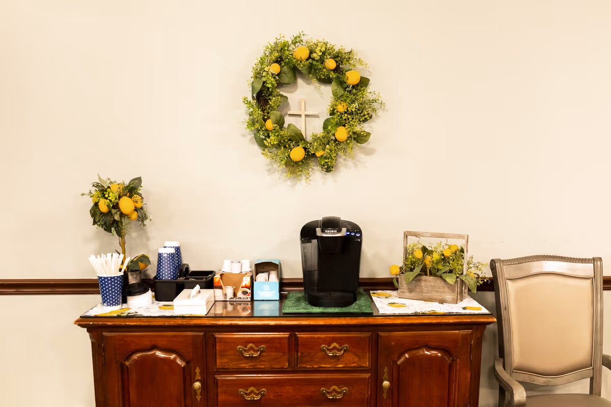 A wooden credenza topped with a Keurig coffee maker, disposable cups and lemon-themed floral decorations under a wreath, with a beige chair to the right.