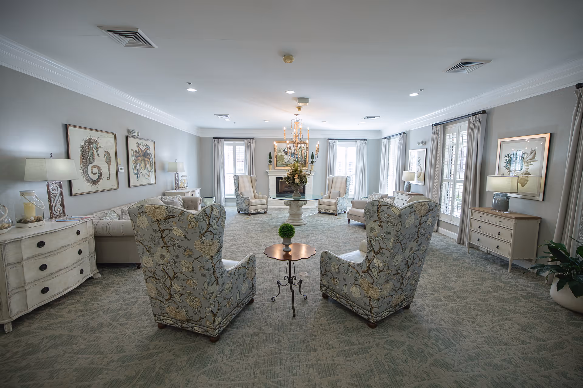 A spacious and elegantly decorated living room with patterned armchairs, a glass-top round table with a floral centerpiece, beige sofas, and multiple lamps on vintage-style dressers. The room features large windows with curtains, framed sea-themed artwork on the walls, and a chandelier hanging from the ceiling.