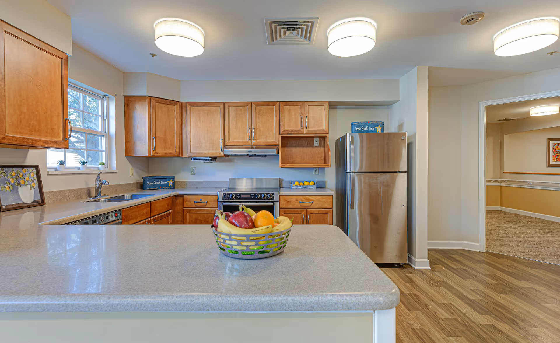 A clean and well-lit kitchen with wooden cabinets, a stainless steel refrigerator, stove, and dishwasher. There is a countertop island in the foreground with a basket of assorted fruits. The kitchen has a window with small potted plants on the sill and opens into a hallway with carpeted flooring.