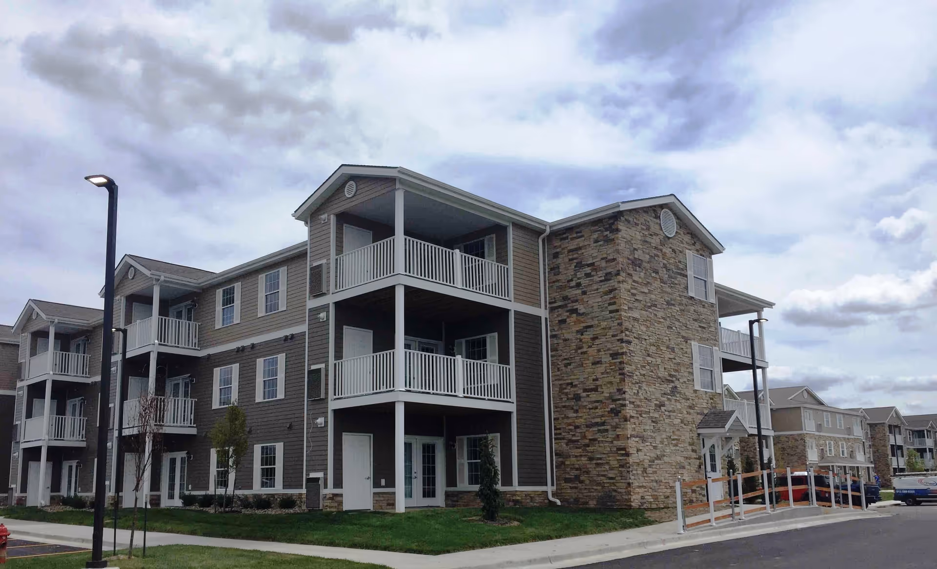 Exterior view of a multi-story residential building with balconies and a stone facade section under a cloudy sky. The building is part of a retirement community with a sidewalk, street lamp, and some small trees in front.