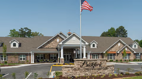 Front exterior view of Bickford of Chesapeake senior living facility with a large American flag on a flagpole in front, a stone sign displaying the facility's name, and a well-maintained driveway and landscaping under a clear blue sky.