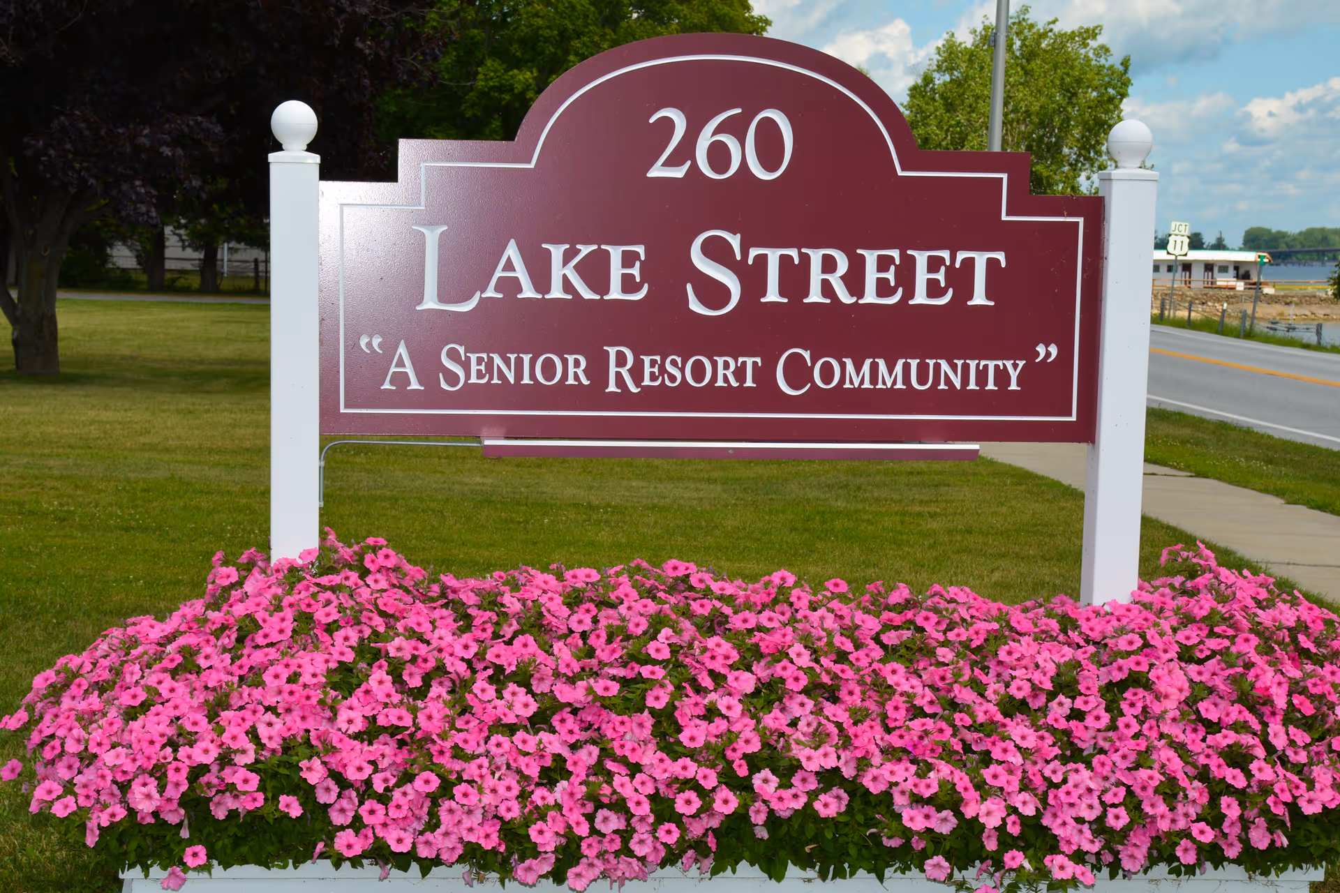 A maroon and white sign reading '260 Lake Street A Senior Resort Community' is displayed outdoors with a bed of vibrant pink flowers in front. The background shows a grassy area, trees, a sidewalk, a road, and a body of water under a partly cloudy sky.