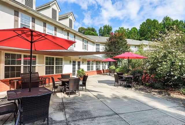 Outdoor patio area at Oaks at Stockbridge with several tables and chairs under red umbrellas, adjacent to a two-story building with white siding and brick accents, surrounded by greenery and trees under a partly cloudy sky.