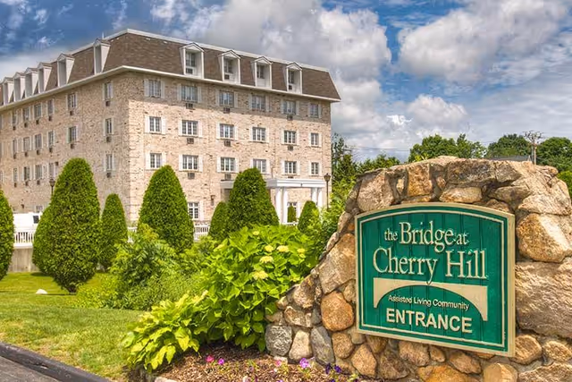 Stone entrance sign for 'The Bridge at Cherry Hill' in front of a landscaped lawn and a multi-story assisted living building under a partly cloudy sky.