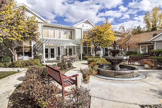 Outdoor courtyard featuring a three-tier fountain, red benches, patio seating, trees, and a two-story residential building under a partly cloudy sky.