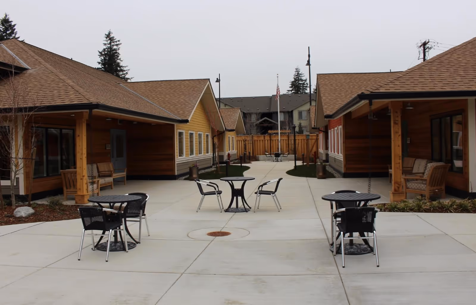 Outdoor courtyard area at Cottages of Renton featuring several small round tables with chairs on a concrete patio, surrounded by single-story buildings with wood siding and pitched roofs. There are benches under covered porches and an American flag on a flagpole in the background.