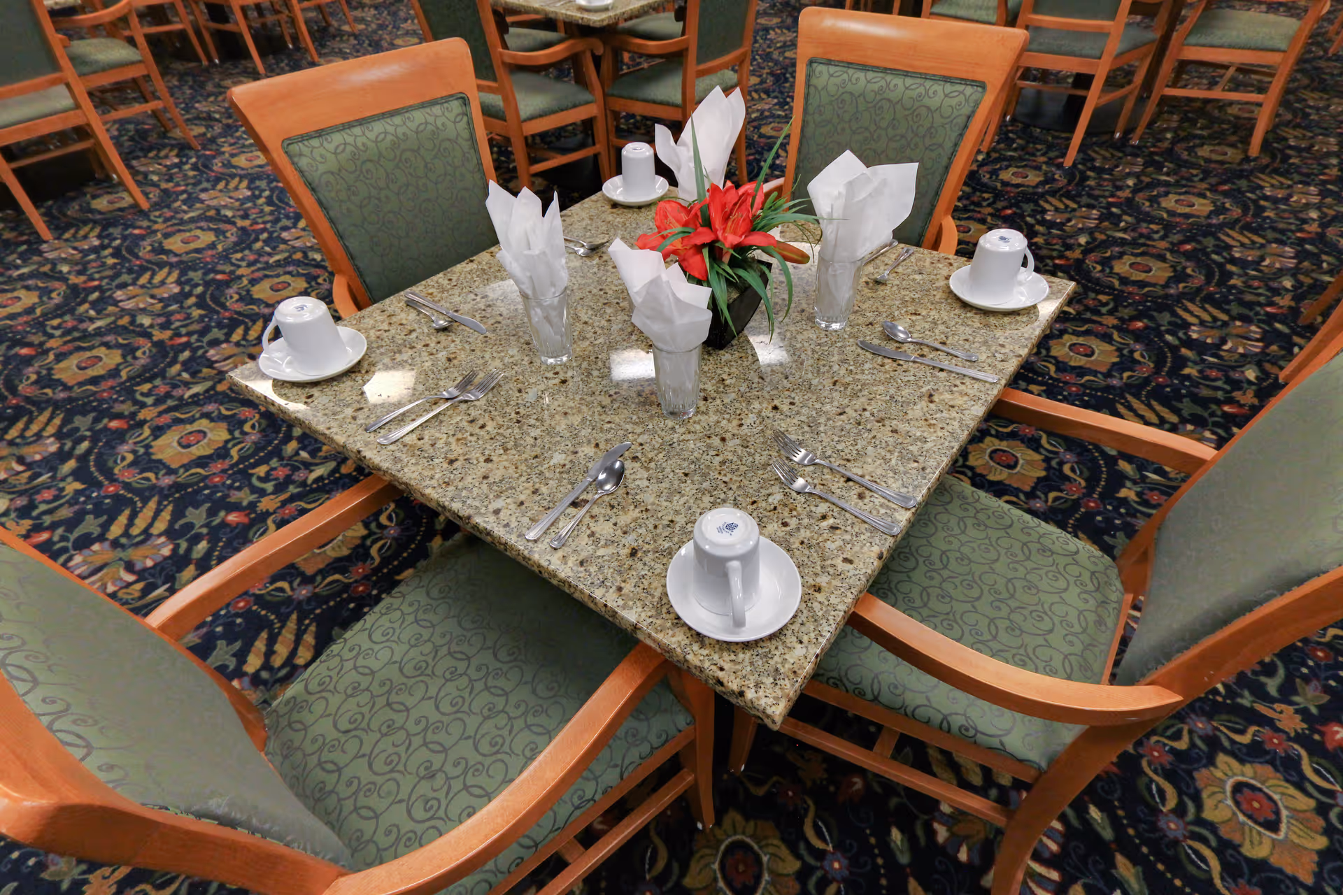 A square dining table set for four with a granite top, green cushioned wooden chairs, white napkins folded in glasses, white coffee cups on saucers, and a red flower centerpiece. The table is placed on a patterned carpet in a dining area with more similar tables and chairs in the background.