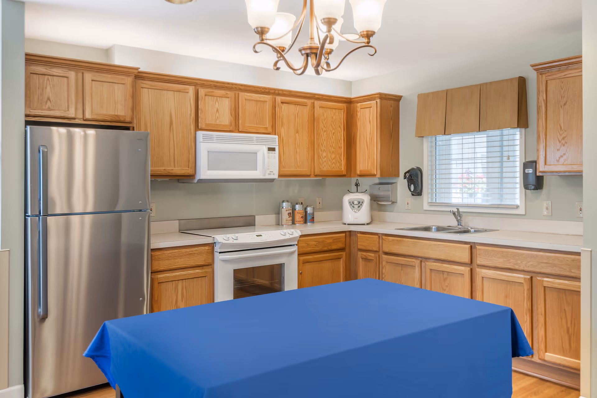 A kitchen with wooden cabinets, a stainless steel refrigerator, a white microwave above a white stove, a sink under a window with blinds and a brown valance, and a table covered with a blue tablecloth in the foreground.