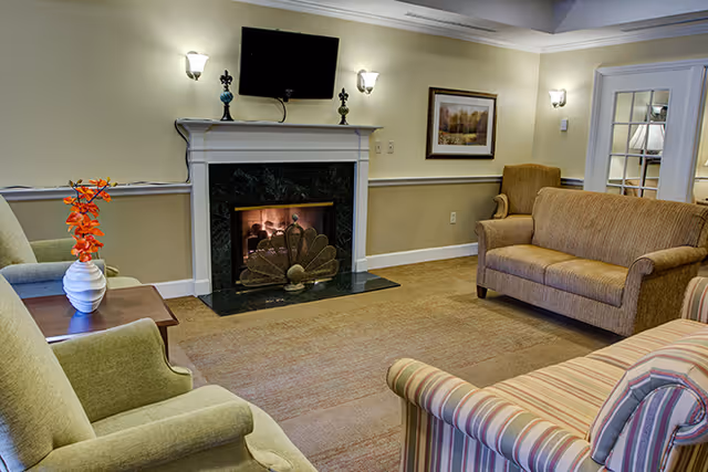 A cozy living room with a lit fireplace featuring a decorative screen, a wall-mounted TV above the mantel, two armchairs, a striped sofa, a brown loveseat, a side table with a white vase holding orange flowers, and a framed picture on the wall.