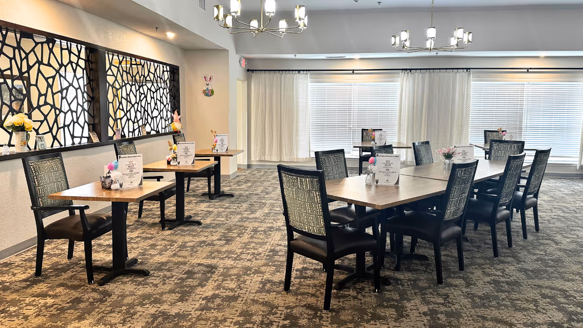 Spacious senior dining room with multiple wooden tables and upholstered chairs, decorative wall partition, chandeliers, and large windows with curtains.