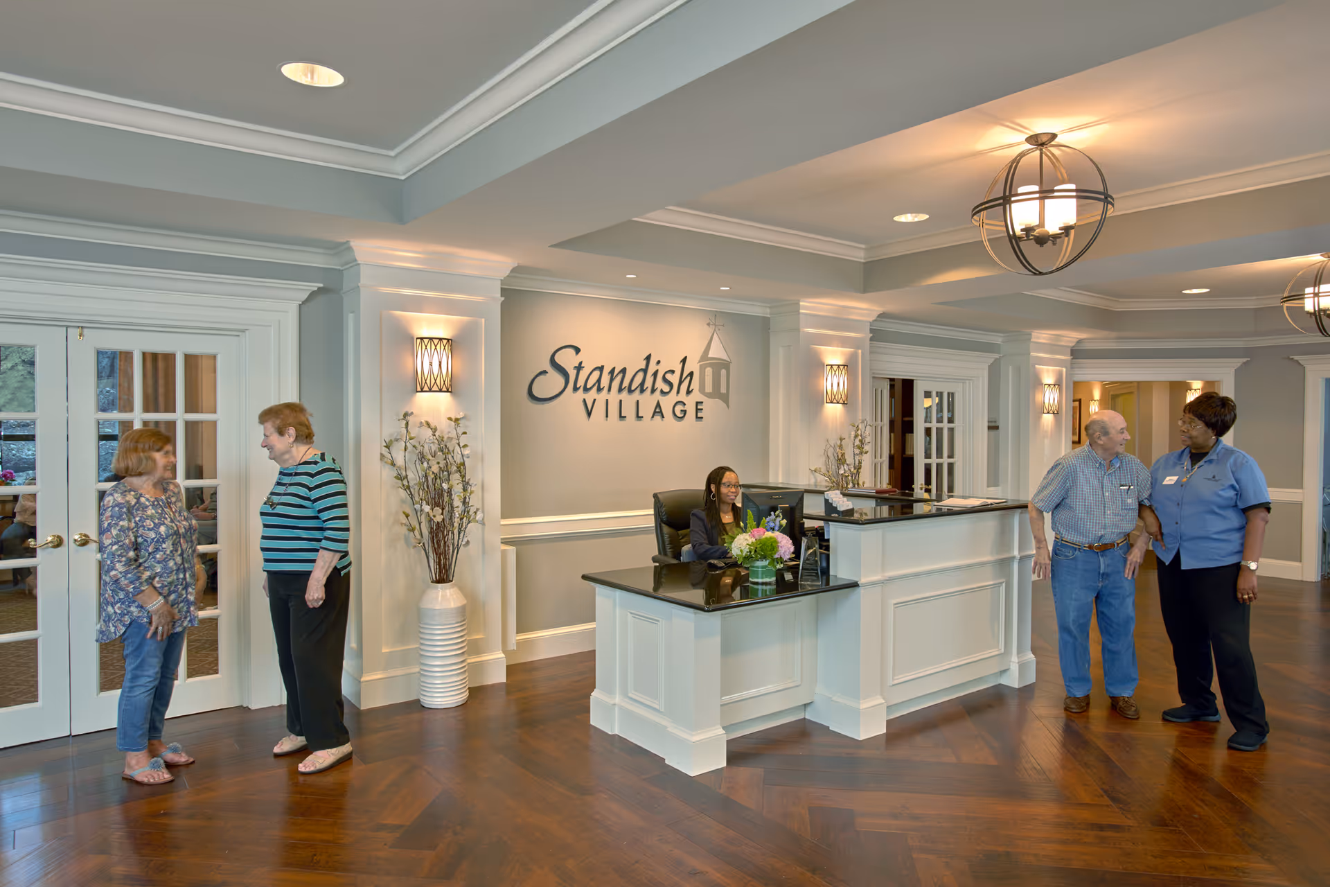 Reception area of Standish Village Assisted Living & Compass Memory Support Community with a receptionist sitting behind a white counter with a black countertop. Two elderly women are conversing near glass double doors on the left, while an elderly man talks with a staff member in a blue uniform on the right. The area has wooden flooring, soft lighting, and decorative plants.