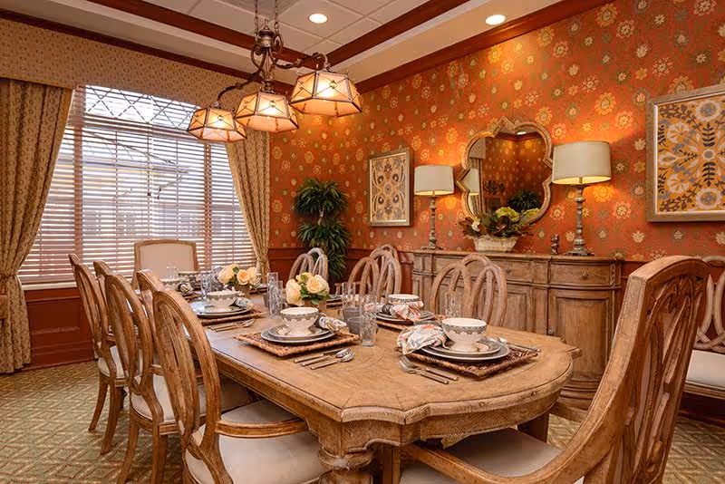 Ornate dining room with a long wooden table set for a meal, upholstered chairs, chandelier, patterned wallpaper, and a sideboard with lamps and mirror.