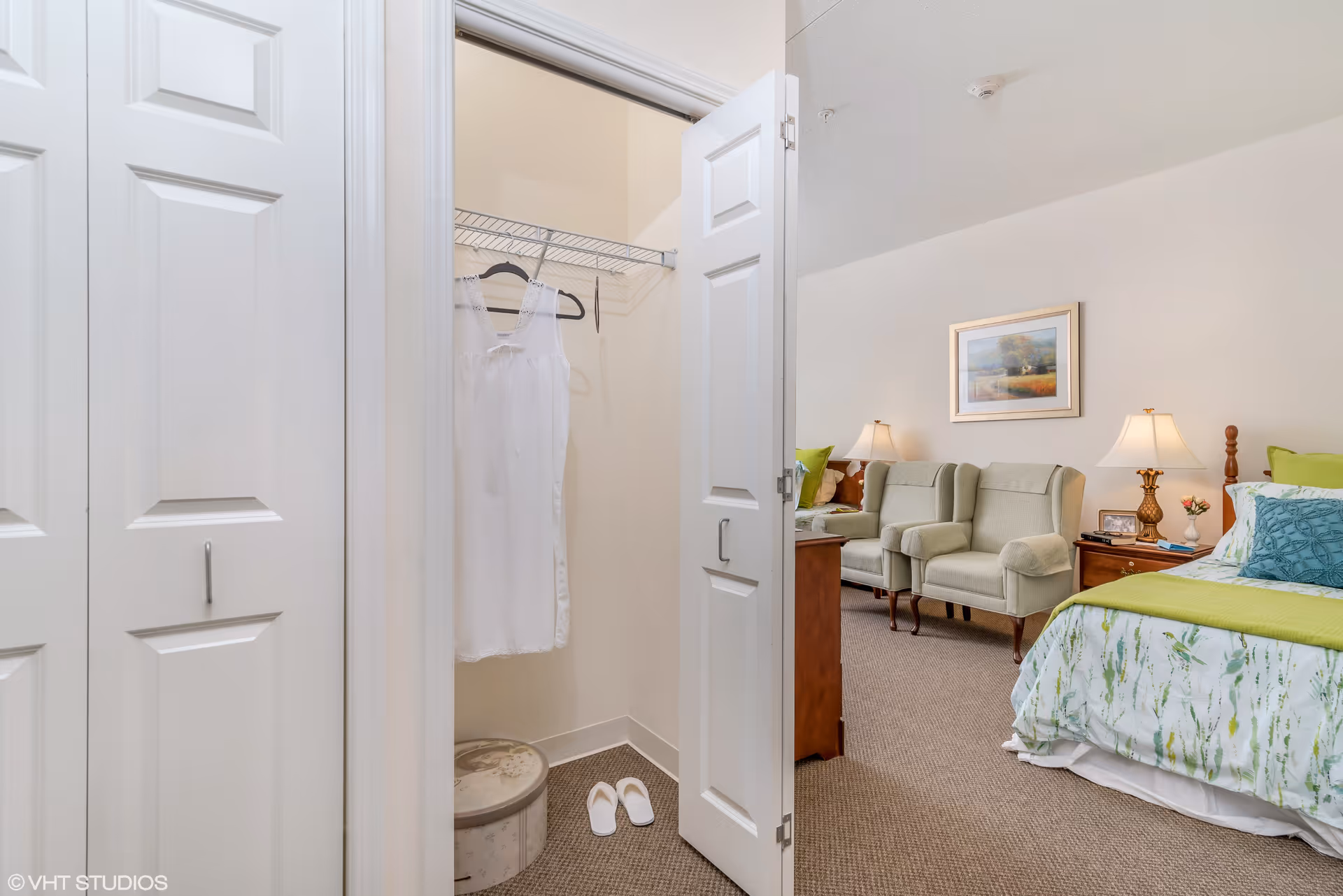 A cozy bedroom with a bed featuring green and white bedding, two green armchairs, a wooden nightstand with a lamp and small decor, and a framed landscape painting on the wall. An open closet door reveals a white dress hanging on a hanger, a round storage box, and a pair of white slippers on the carpeted floor.