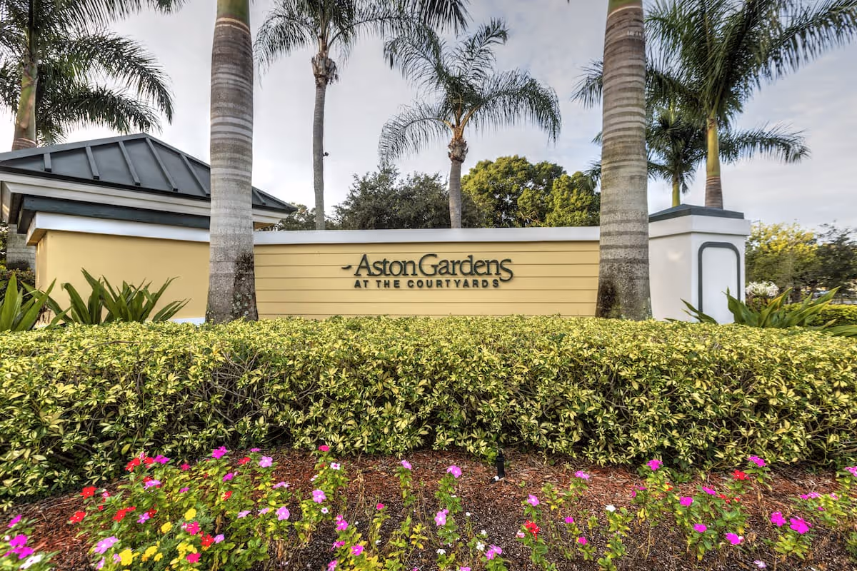 Entrance sign for Aston Gardens At The Courtyards surrounded by palm trees, green shrubs, and colorful flowers under a partly cloudy sky.