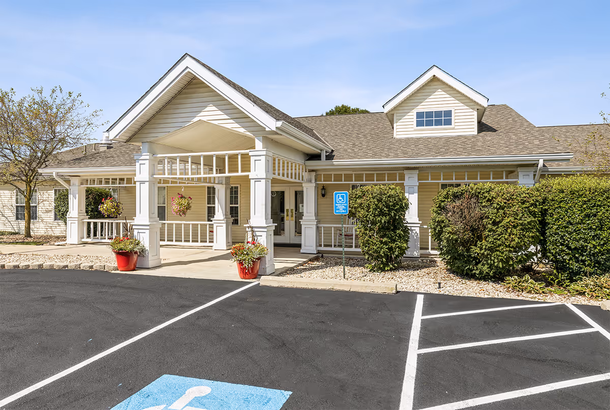Exterior view of a senior living facility entrance with a covered porch supported by white columns, hanging flower baskets, and a handicapped parking space in front. The building has beige siding and a gabled roof under a clear blue sky.