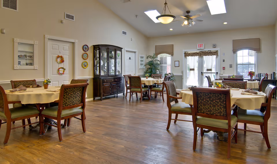 A bright dining room in a senior living facility with several round tables covered with beige tablecloths, each surrounded by wooden chairs with patterned upholstery. The room has wooden flooring, a ceiling fan with lights, and large windows with curtains allowing natural light to enter. There is a dark wooden china cabinet against one wall and decorative plates hanging nearby.