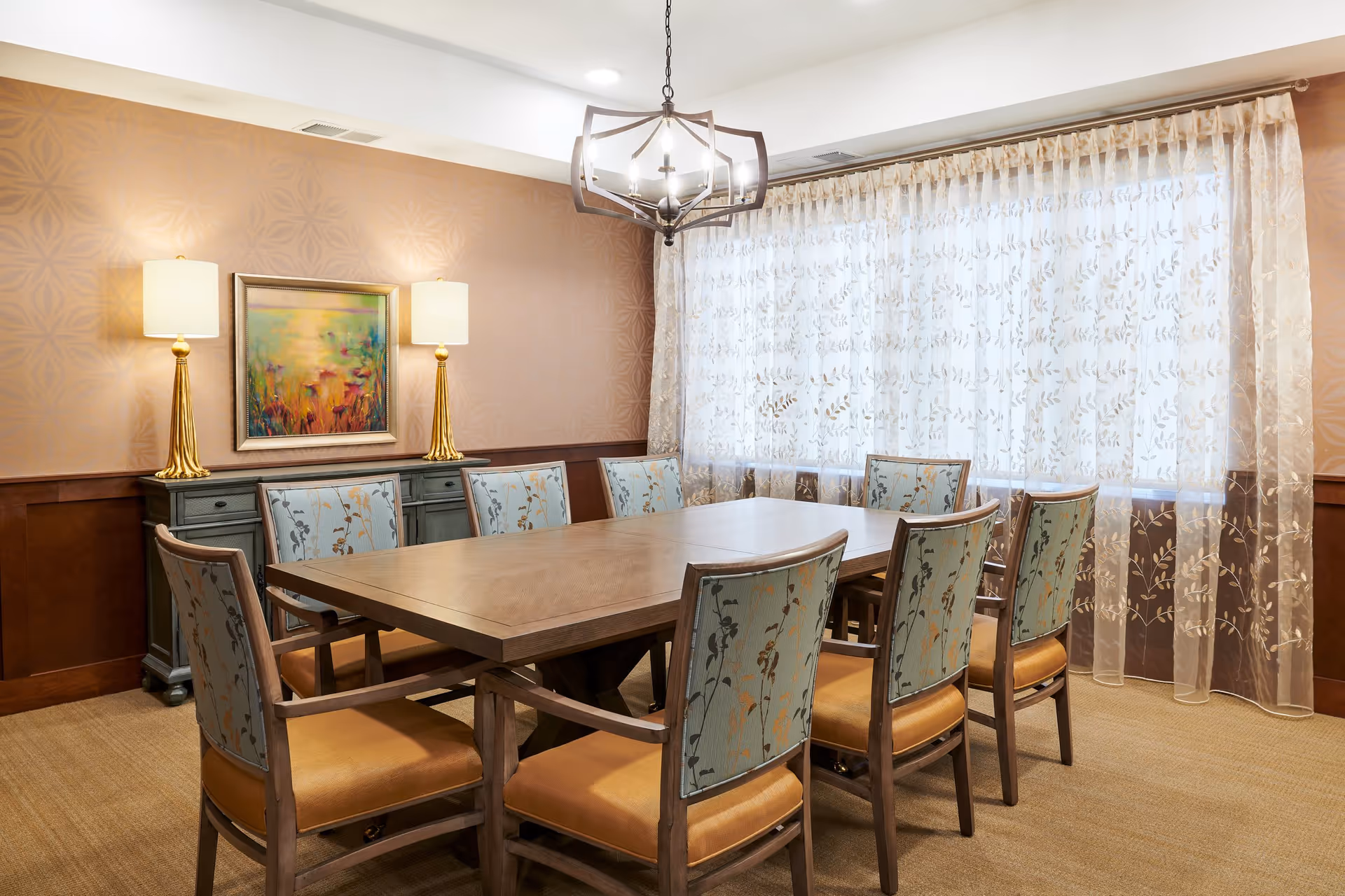 A well-lit dining room with a large wooden table surrounded by eight upholstered chairs with floral patterned backs and tan seats. The room features a decorative chandelier hanging above the table, sheer curtains with a leaf pattern covering a large window, two table lamps with gold bases on a sideboard, and a colorful framed painting on the wall.