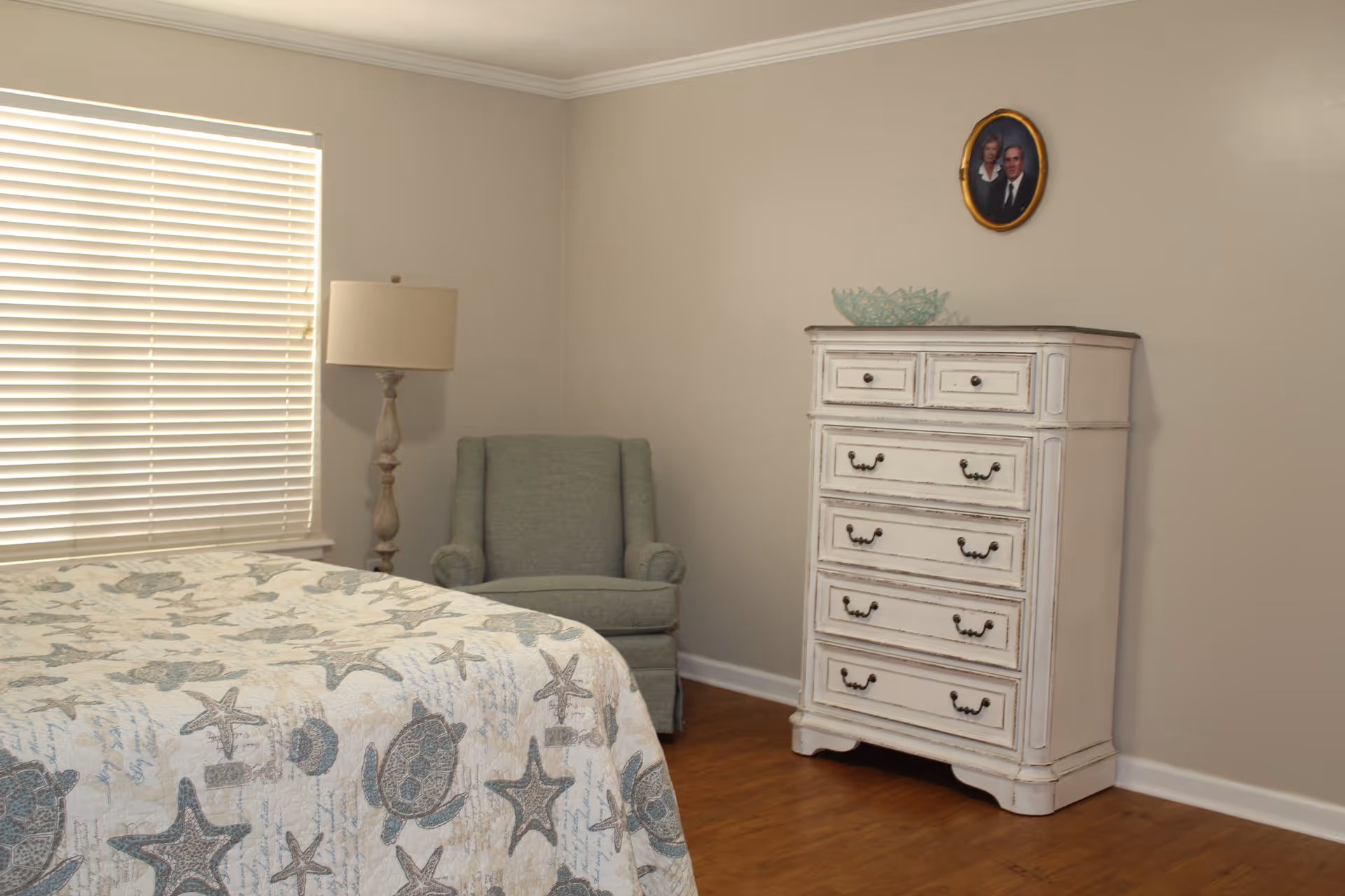 A bedroom with a bed covered in a quilt featuring sea turtles and starfish patterns, a light green upholstered armchair, a tall white distressed dresser with multiple drawers, a floor lamp with a beige shade, and a small oval framed portrait hanging on the beige wall above the dresser. The room has wooden flooring and a window with closed blinds.