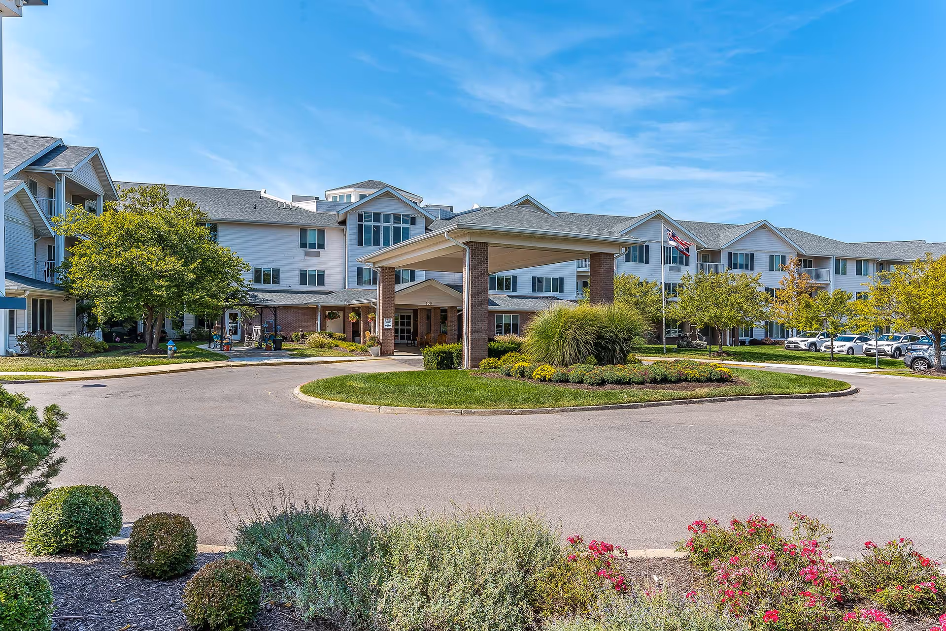 Exterior view of Solstice Senior Living at Columbia, showing a large three-story building with white siding and gray roofing. The entrance features a covered driveway with brick pillars, surrounded by landscaped greenery, bushes, and flowers. Several cars are parked along the driveway, and an American flag is visible near the entrance under a clear blue sky.