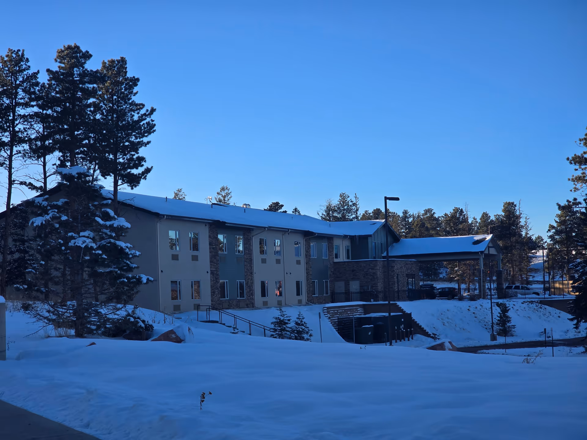 Exterior view of a two-story senior living facility building named Forest Ridge Senior Living, surrounded by snow-covered ground and pine trees under a clear blue sky.