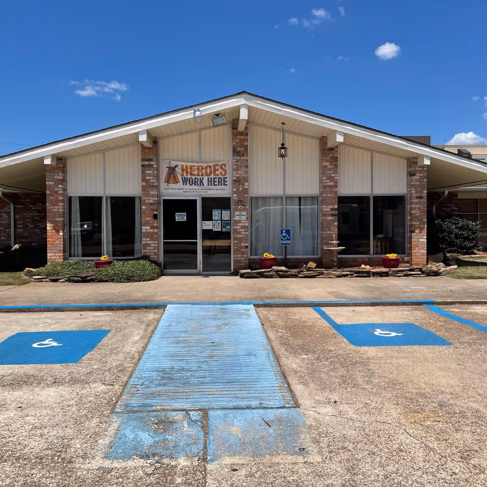 Front exterior view of Claiborne Healthcare Center building with a brick facade, large windows, and a sign above the entrance that reads 'HEROES WORK HERE'. There are two handicap parking spaces and a blue wheelchair accessible ramp leading to the entrance. The sky is clear and blue with a few clouds.