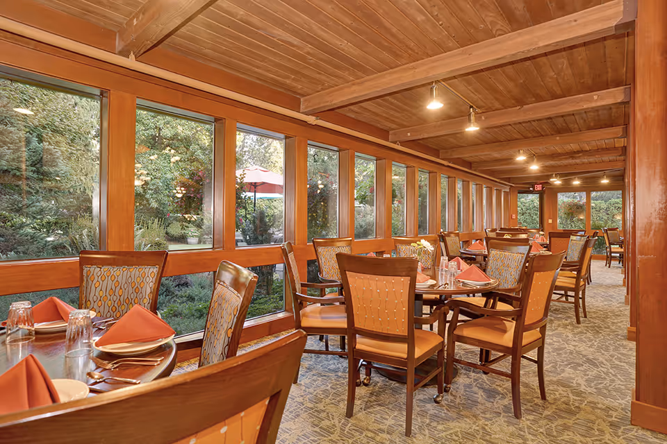 Dining room with multiple round tables set with plates, glasses, and folded orange napkins. The room has large windows along one side showing a garden with greenery and an umbrella outside. The interior features wooden beams and ceiling with carpeted flooring.