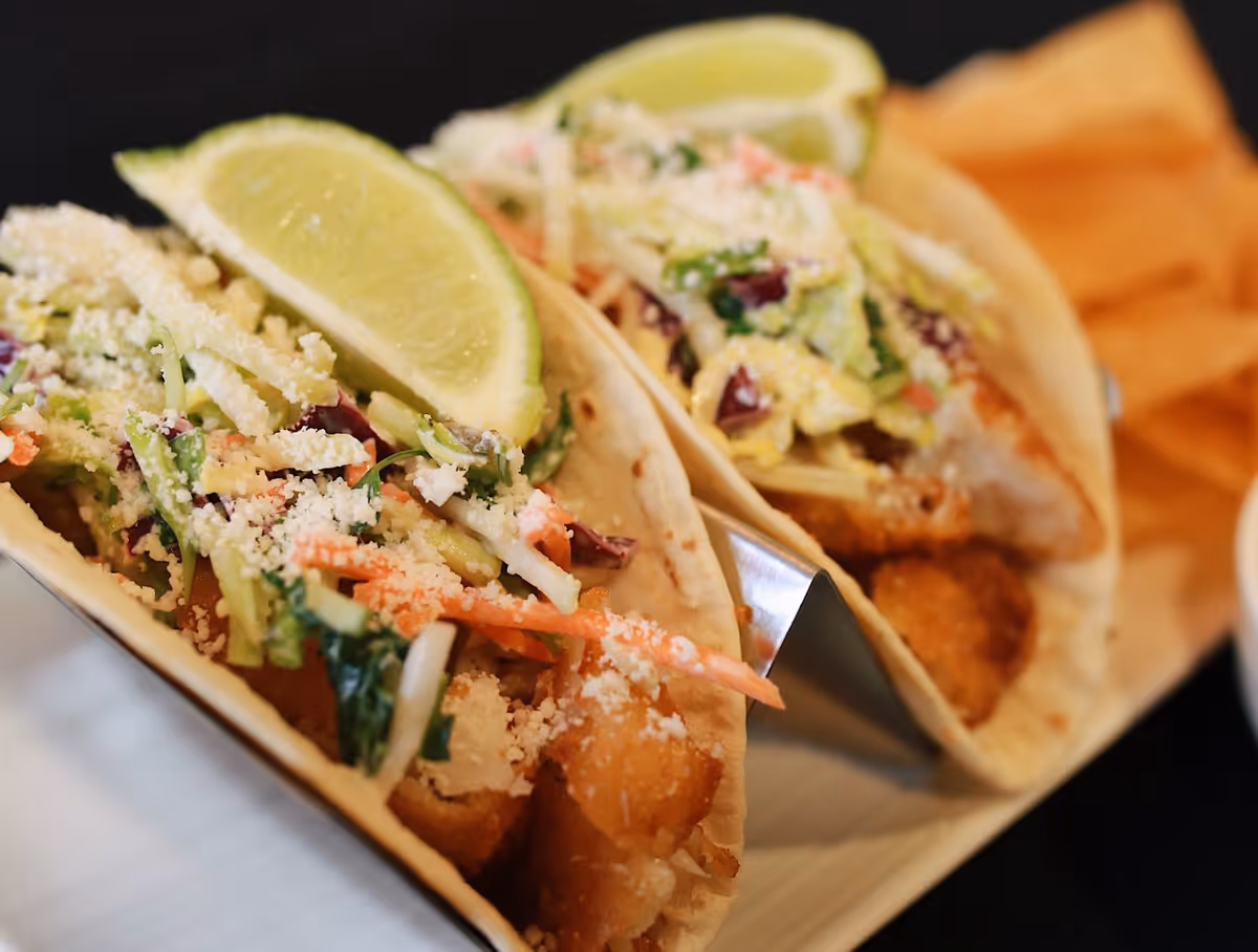 Close-up of two fish tacos topped with shredded slaw, crumbled cheese, and lime wedges.