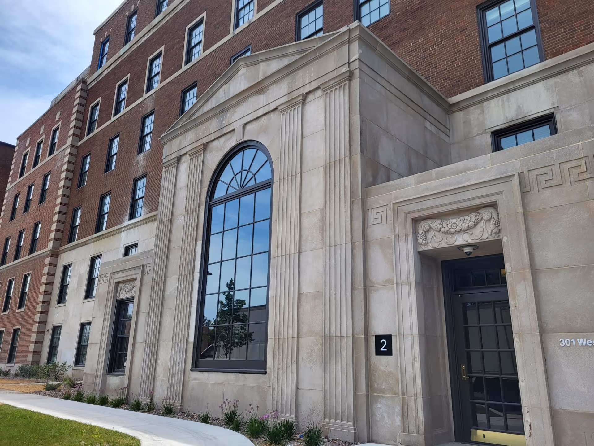 Exterior view of a multi-story brick and stone building with large arched windows and decorative stonework around the entrance. There is a sidewalk and some landscaping with grass and plants in front of the building.