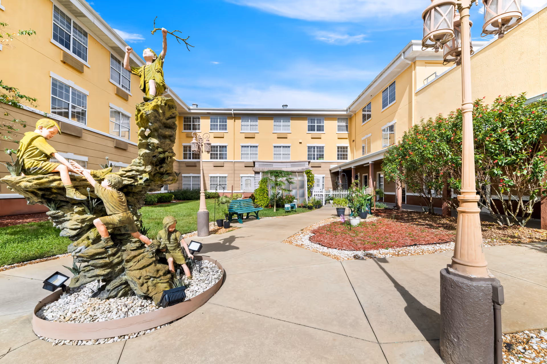 Outdoor courtyard area of a senior living facility with a sculpture of children playing on a rock formation, surrounded by a paved walkway, green benches, bushes, and a three-story yellow building in the background under a blue sky.