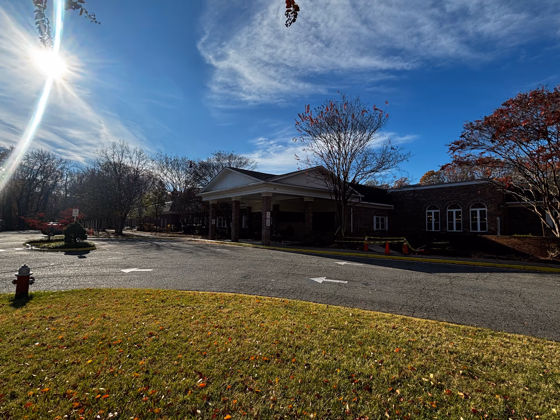 Exterior view of a brick building with a covered entrance, surrounded by trees with autumn foliage under a bright blue sky with some clouds. There is a grassy area with scattered fallen leaves in the foreground and a paved driveway with directional arrows leading to the entrance.