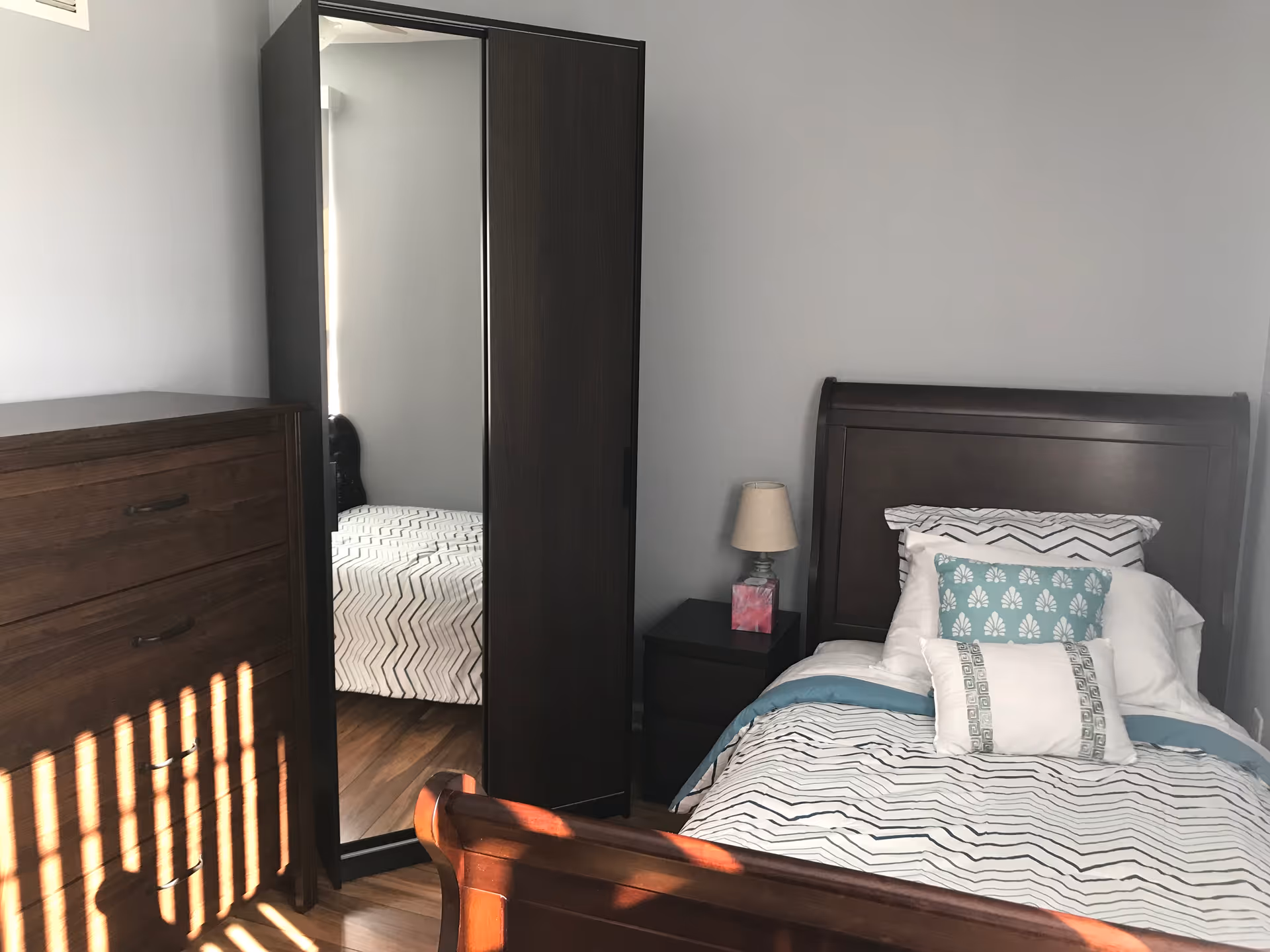 Small bedroom with a single bed, nightstand and lamp, wooden dresser, and a mirrored wardrobe against a gray wall.