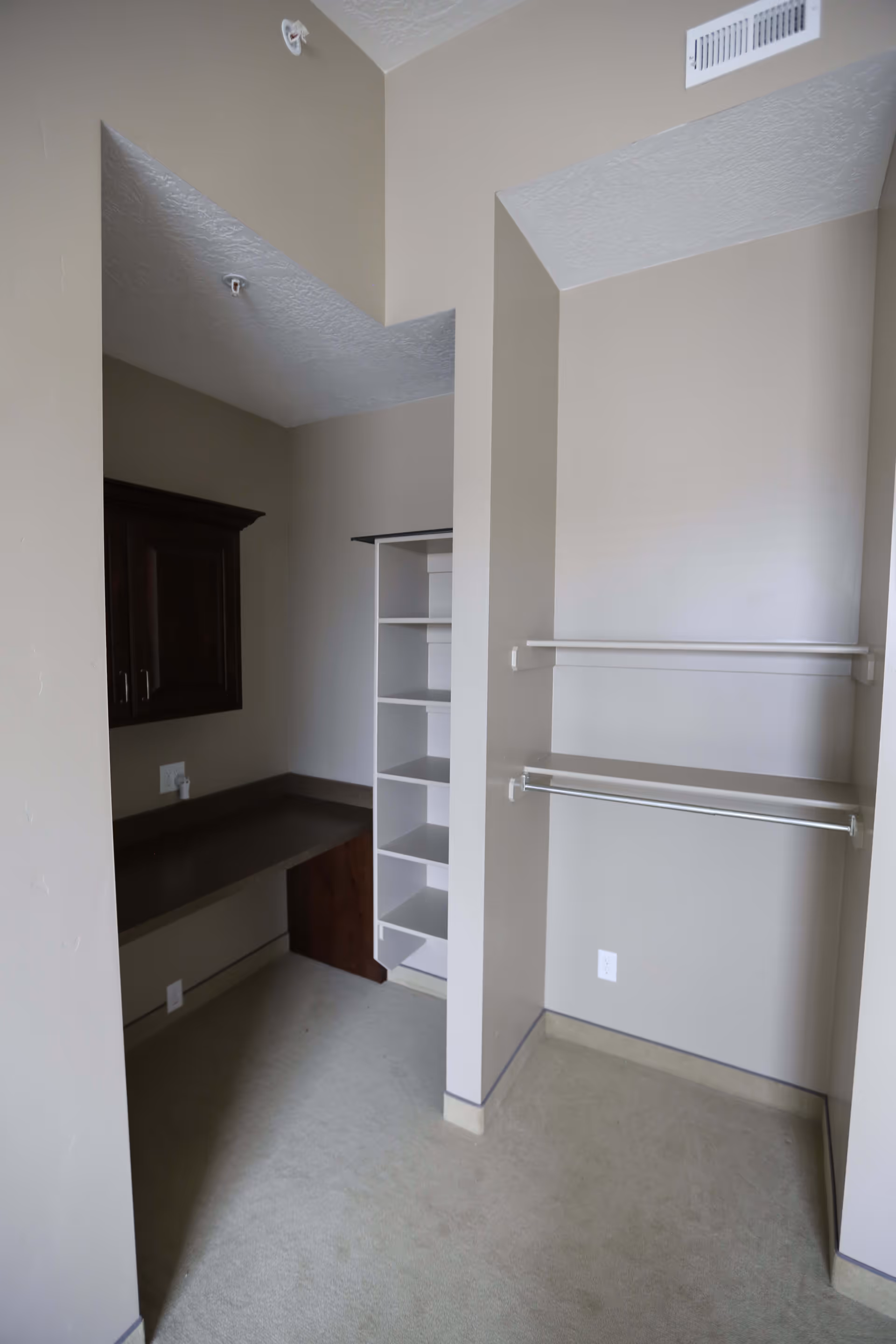 An empty walk-in closet area with beige walls and carpeted floor. The closet features white shelving units, a hanging rod, and a built-in dark wood cabinet with a countertop workspace underneath.
