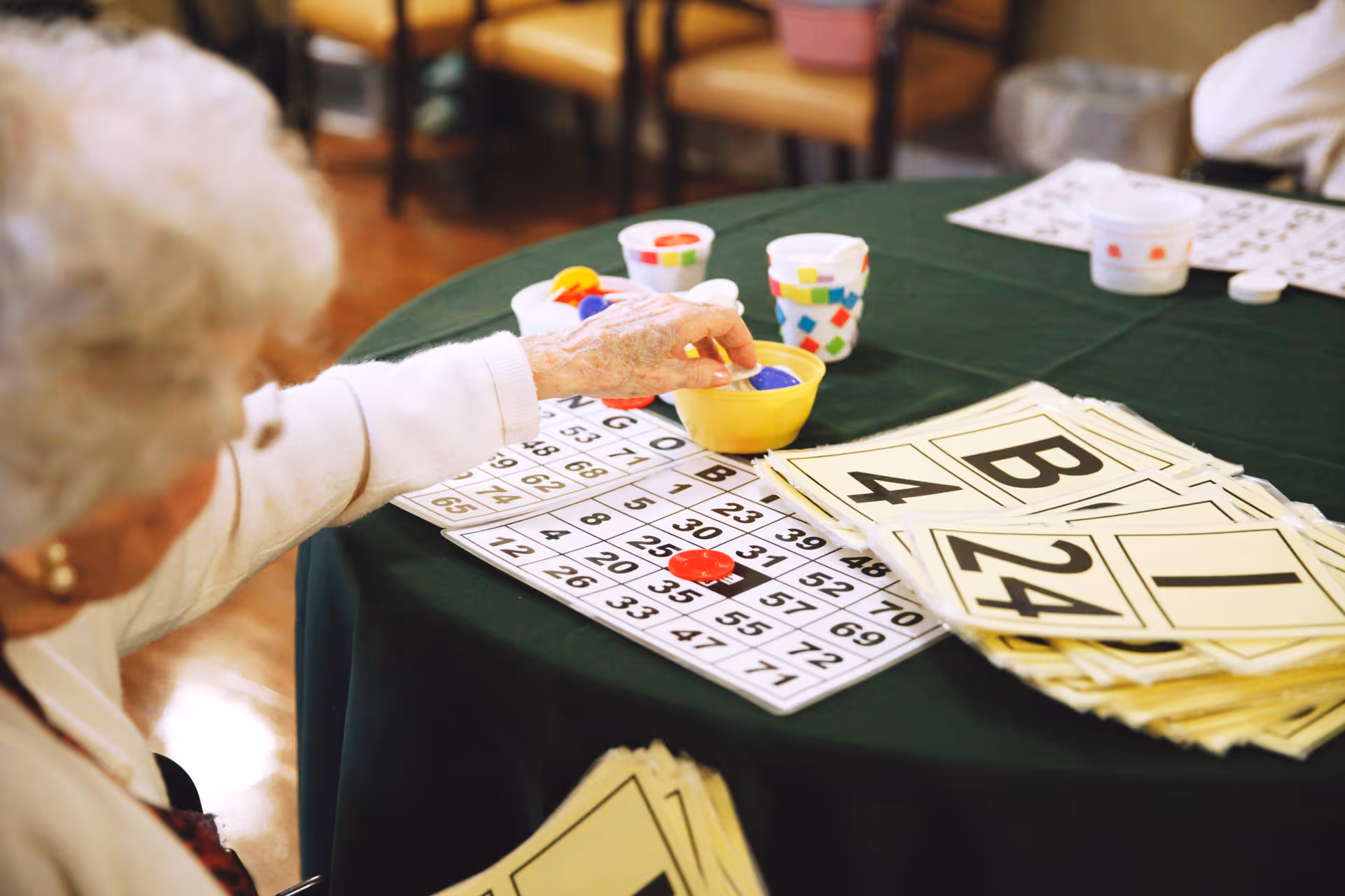 An elderly person playing bingo at a table covered with a green tablecloth, placing a blue marker on a bingo card. Several bingo cards and colorful cups with markers are also on the table.