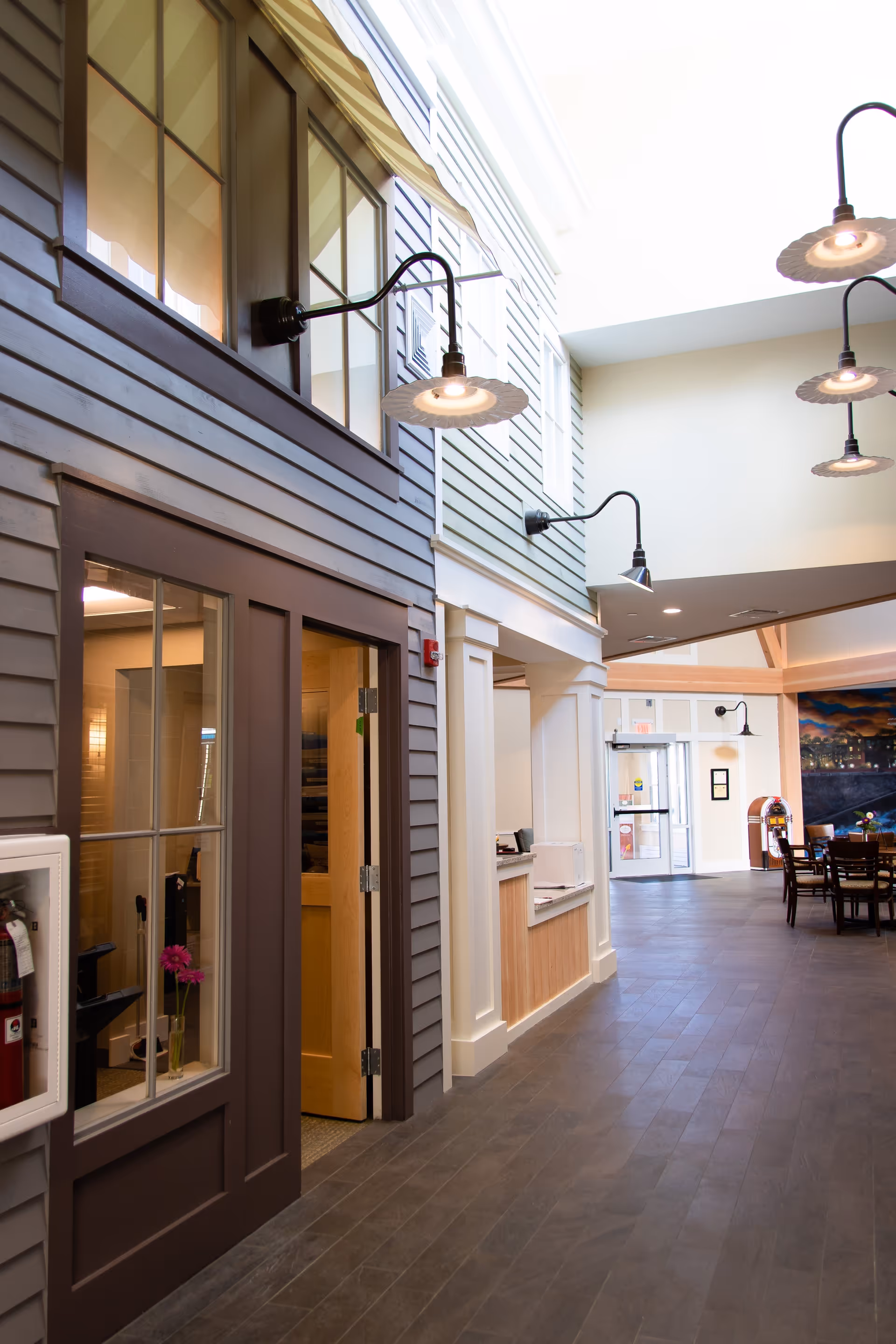Interior hallway of a senior living facility with high ceilings, wall-mounted lamps, and a seating area with tables and chairs in the background. The walls have a house-like design with windows and doors, and there is a fire extinguisher mounted on the wall.