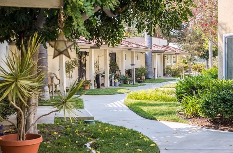 A sunny outdoor walkway at Waterford Terrace Retirement Community lined with single-story residential units. The path curves through well-maintained green lawns and landscaped bushes, with potted plants and trees providing shade and decoration.