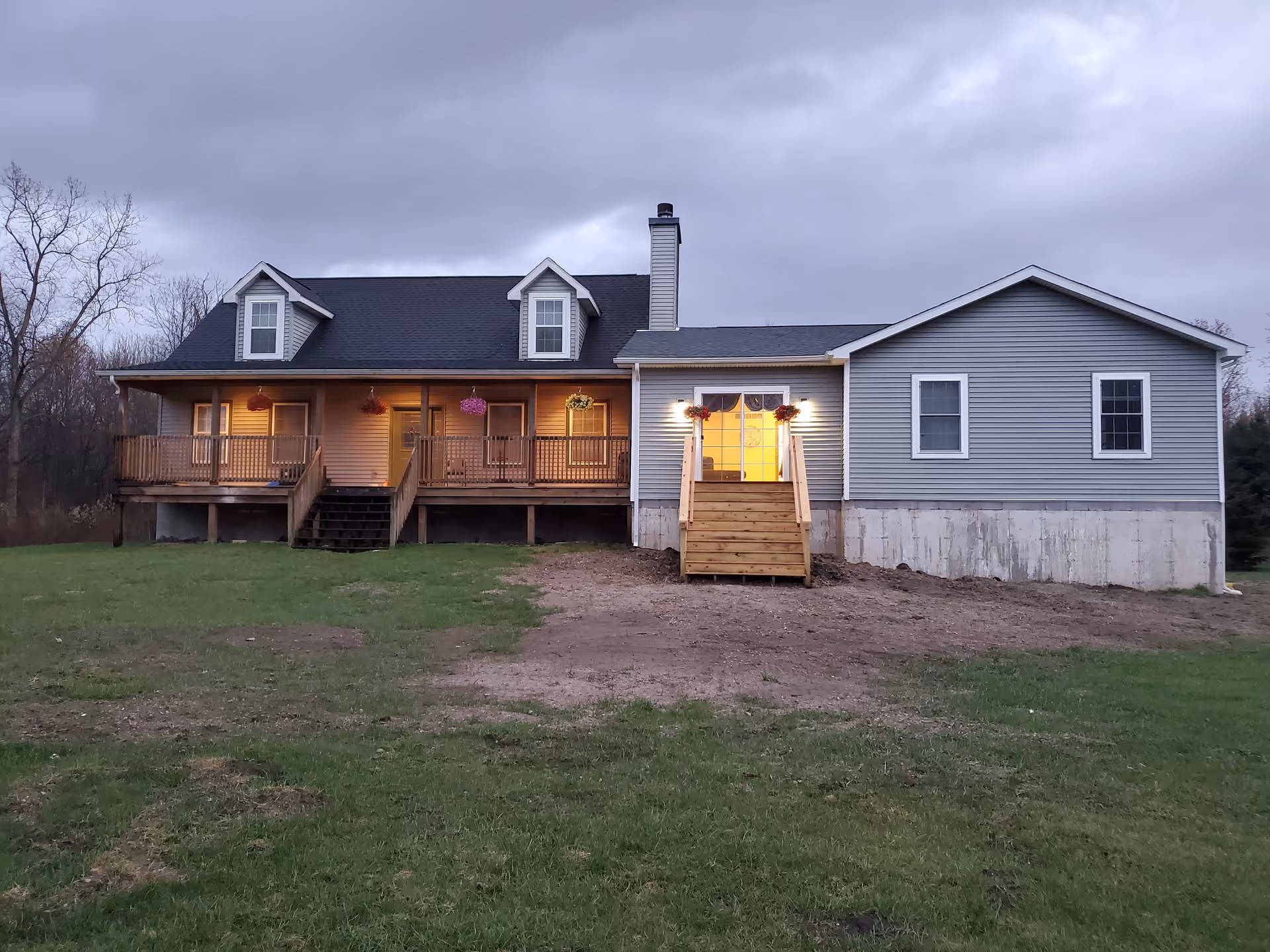 Exterior view of a single-story house with a porch and stairs leading to a door illuminated by lights. The house has gray siding, a black roof with two dormer windows, and a chimney. The surrounding area includes a grassy lawn and some bare patches of soil under a cloudy sky.