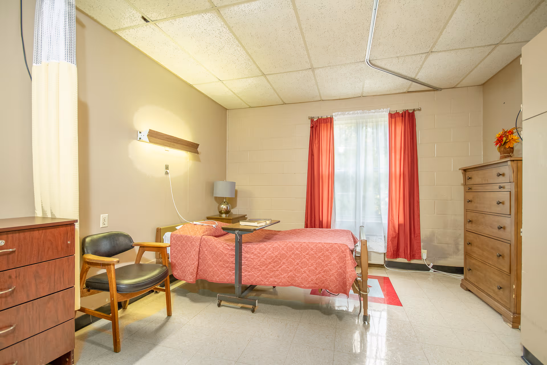 A simple nursing facility bedroom with a single bed covered in a red quilt, a wooden dresser, a wooden chair with black cushion, a bedside table with a lamp, and a window with red curtains allowing natural light to enter.