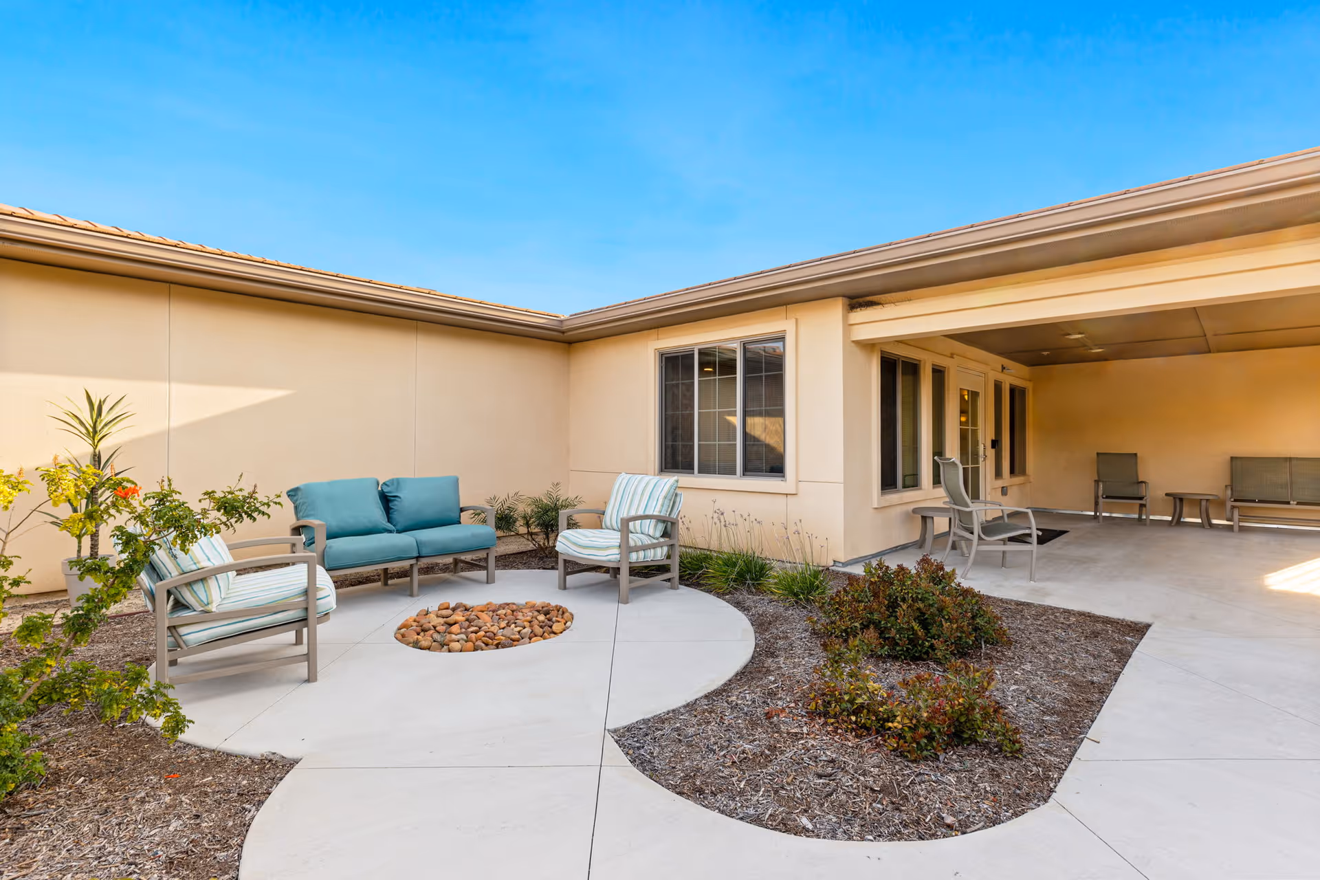 Outdoor courtyard area with seating including a teal cushioned loveseat and two striped cushioned chairs around a circular fire pit filled with rocks. There are plants and mulch beds along the edges and a covered patio area with additional chairs and tables. The building walls are beige with windows and a clear blue sky overhead.