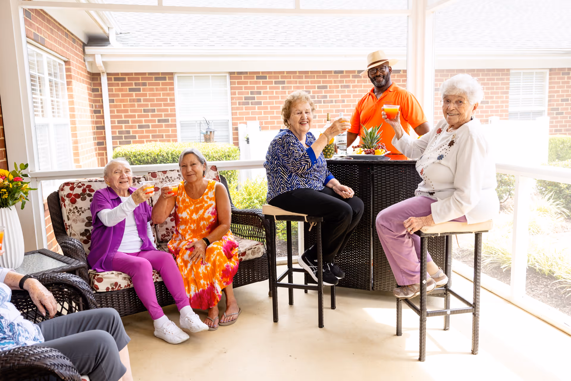 A group of elderly people and a staff member enjoying drinks together on a covered patio with wicker furniture and a brick building in the background. They are smiling and raising their glasses in a toast.