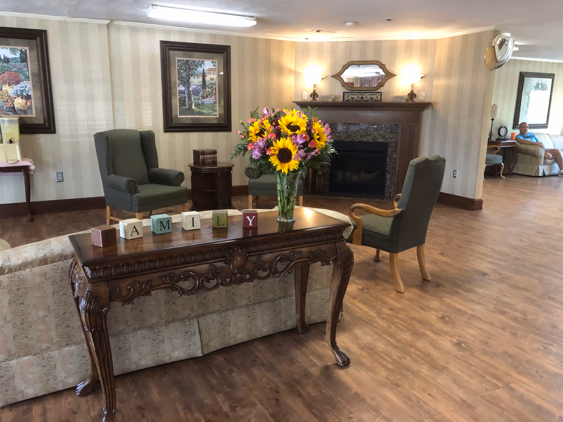 A cozy living room area with wooden flooring, a decorative wooden table with a vase of sunflowers and blocks spelling 'FAMILY'. There are green upholstered armchairs, a fireplace with a mirror and wall sconces above it, framed paintings on the walls, and a person sitting on a couch in the background.
