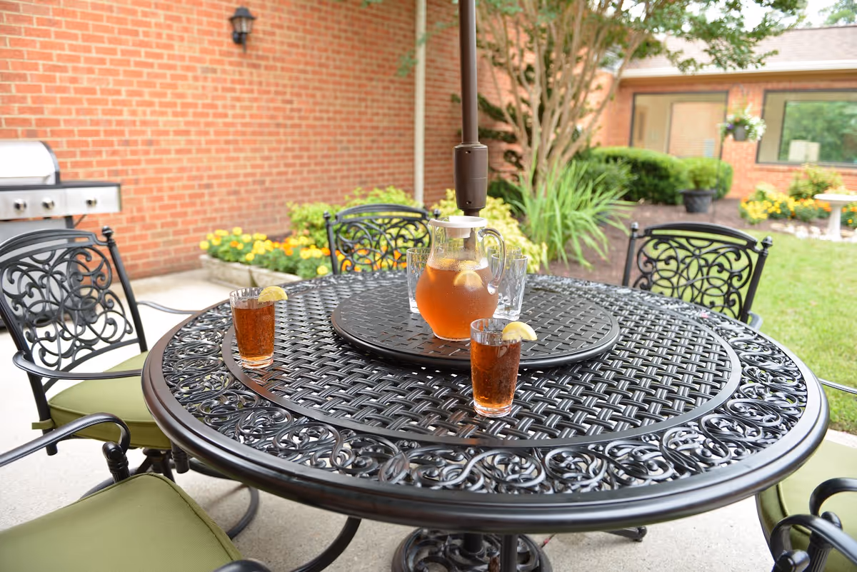Outdoor patio with an ornate metal table holding a pitcher of iced tea and glasses, surrounded by chairs and landscaping beside a brick building.