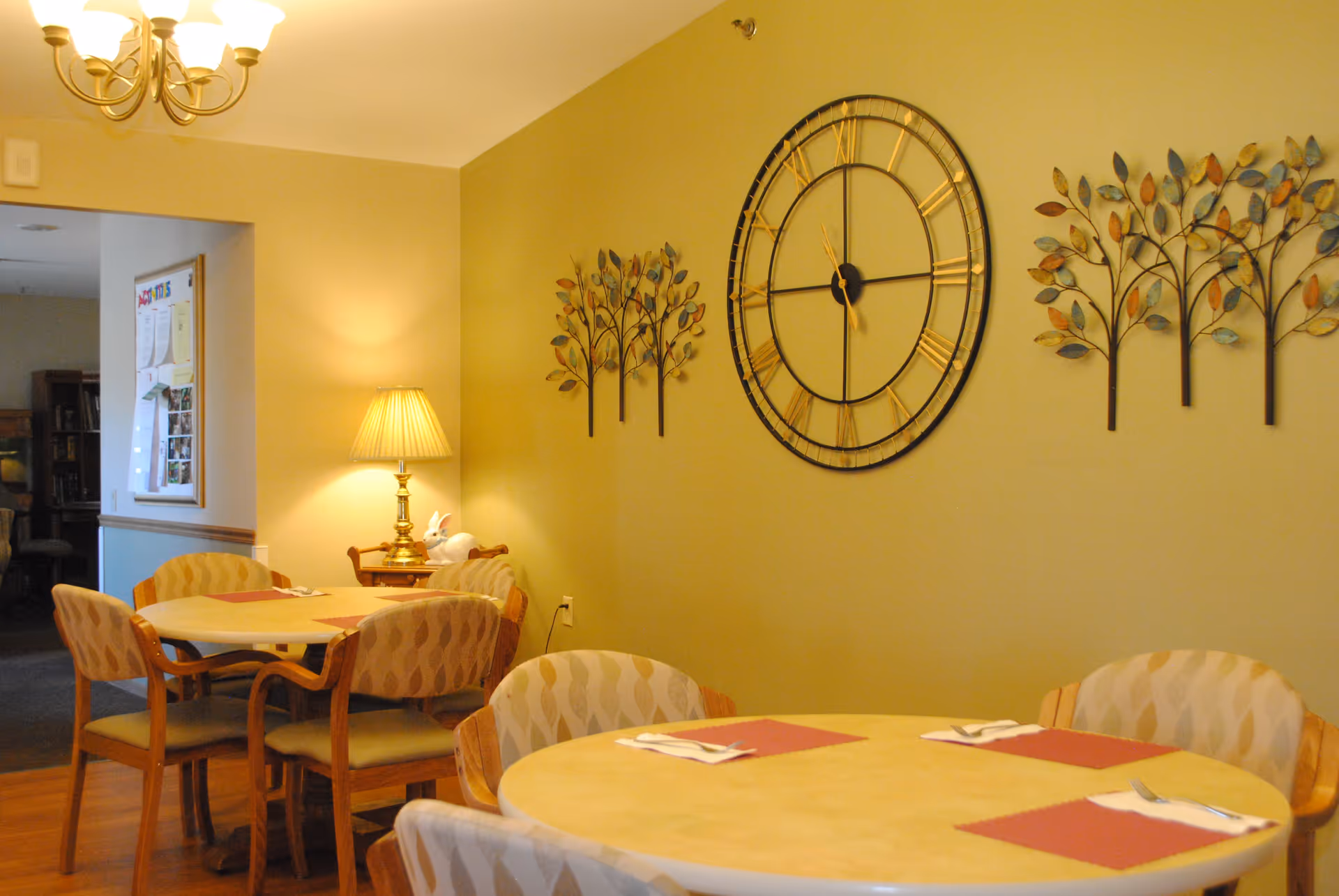 A cozy dining area with round tables set with placemats and silverware. The room features patterned chairs, a decorative wall clock, and metal wall art shaped like trees. A table lamp provides warm lighting, and a chandelier hangs from the ceiling.
