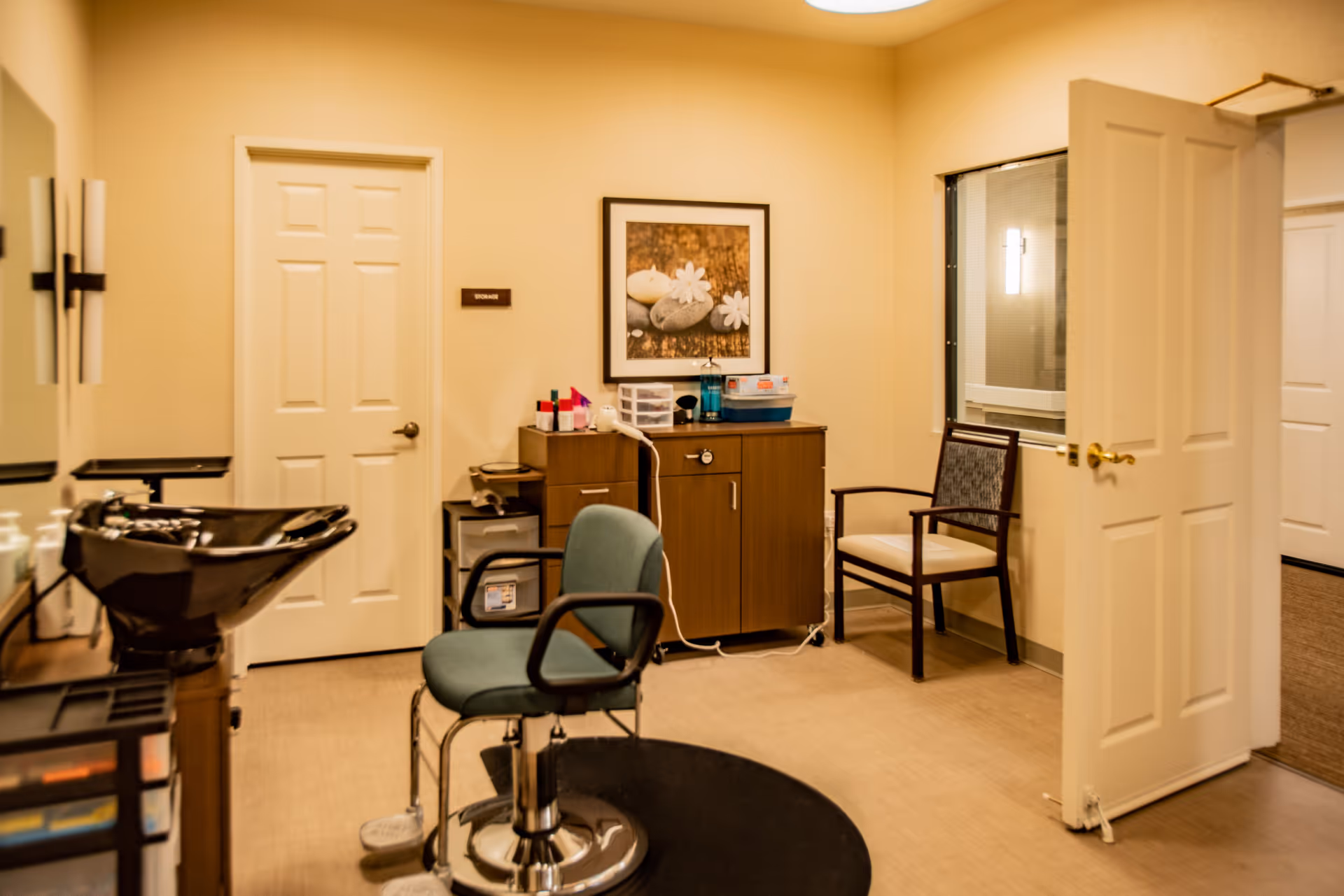 Interior of a hair salon area within a senior living facility, featuring a salon chair, a black hair washing sink, a wooden cabinet with various hair care products, a framed picture on the wall, and two doors, one labeled 'Storage'.