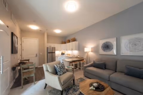 Open-plan living area with a sofa, armchair, coffee table, and a small kitchen and dining nook in the background.
