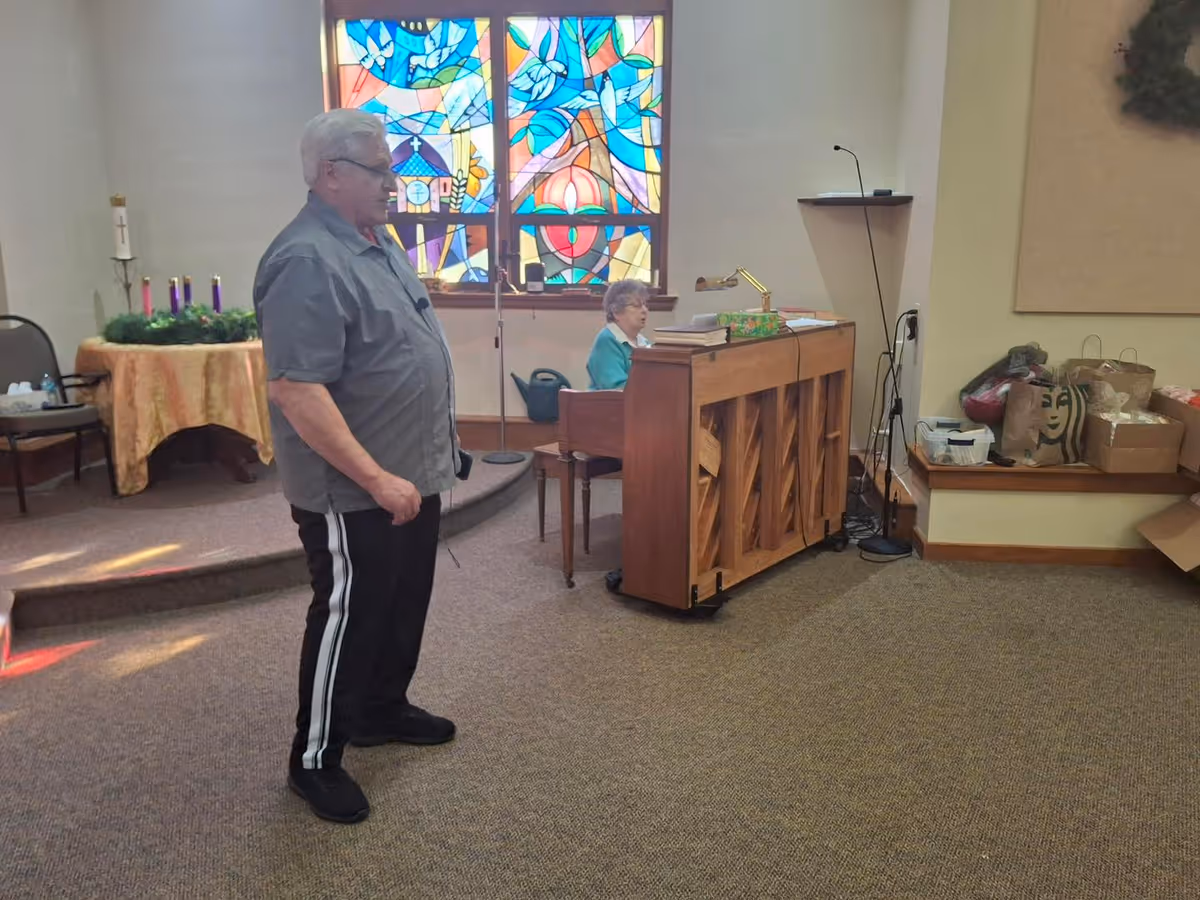 A man stands near a piano while a woman plays it in a chapel-like interior with a colorful stained-glass window.